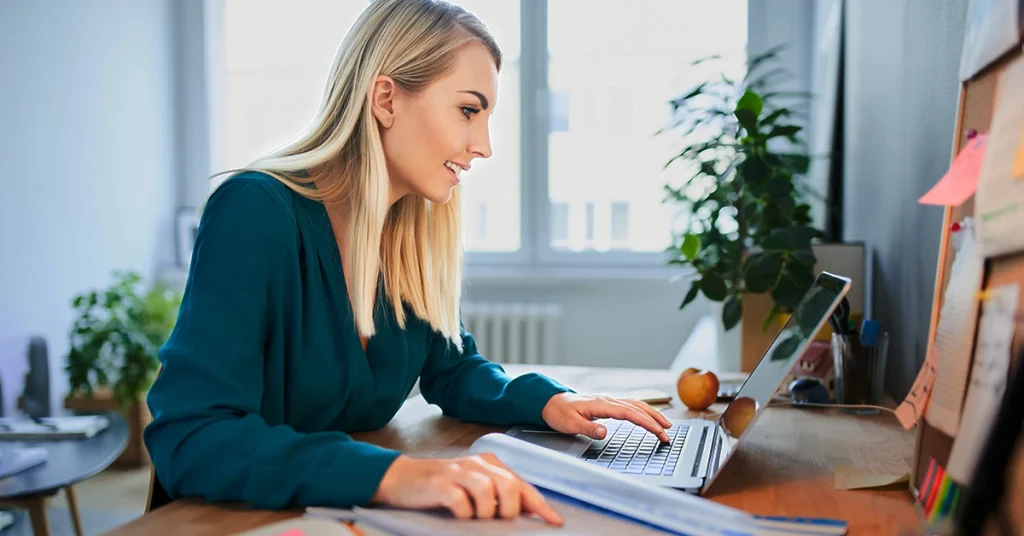 Woman working on a laptop at a desk with papers and an apple, representing article on 8 fun facts about Leap Day.