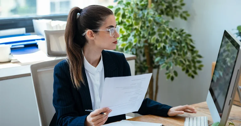 woman reviewing documents while working on a desktop computer in an office, representing Internal Control: Objectives and Components.