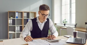 Focused accountant working with financial documents, calculator, and laptop in a bright office, symbolizing the AICPA’s preparations for Canadian CPA program expansion.