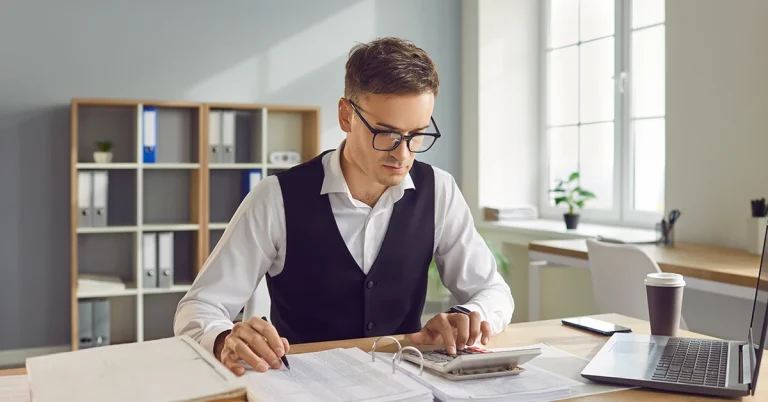 Focused accountant working with financial documents, calculator, and laptop in a bright office, symbolizing the AICPA’s preparations for Canadian CPA program expansion.