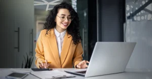 An accountant in a yellow suit jacket works on a laptop at her desk.