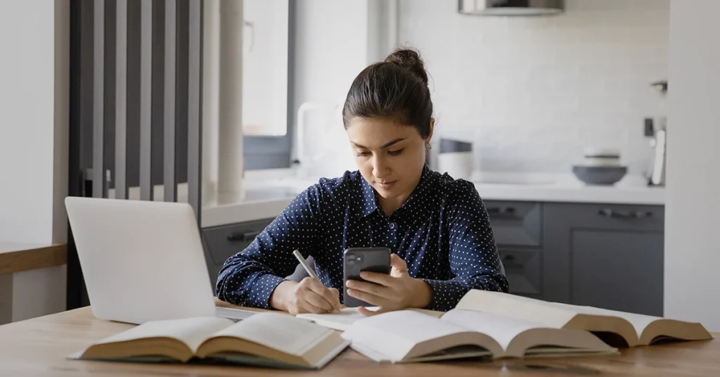 Student using the best CPA review app on their phone while taking notes, surrounded by books.