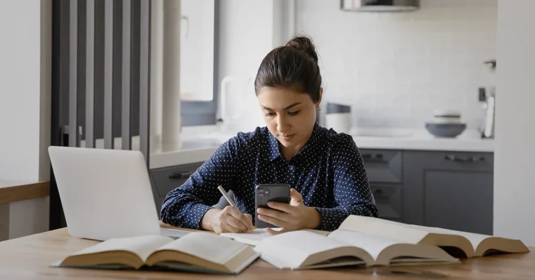 Student using the best CPA review app on their phone while taking notes, surrounded by books.